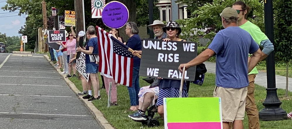 A group of protestors stand along a street in Mantua. They hold signs and American Flags.