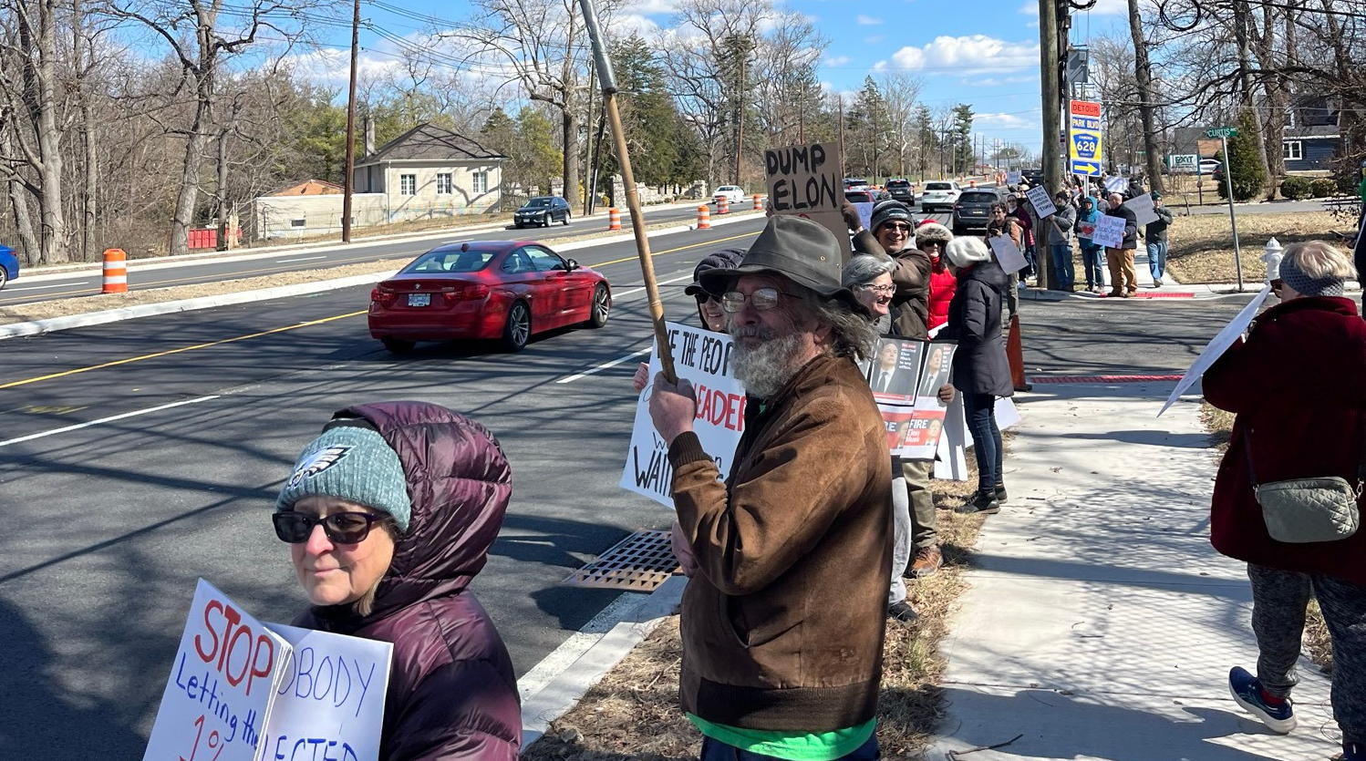 People protest along Route 70 in Cherry Hill. We have people with signs and flags on the left side of the picture as a car drives down the road.
