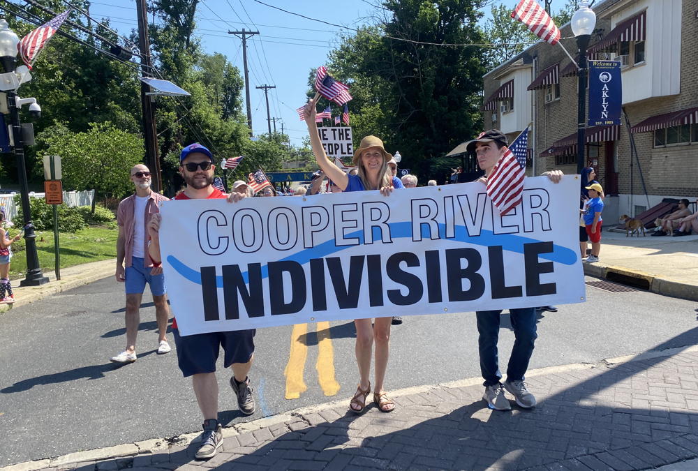 Three people walk up the street in Oaklyn as part of the 4th of July parade. They hold a banner that says "Cooper River Indivisible"