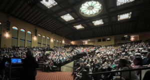 A picture of the audience for the Scottish Rite Auditorium. We see the beautiful stained glass lights on the ceiling and a full house in the crowd