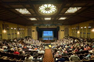 A picture taken from the back of the auditorium showing a packed house and speakers on the front stage