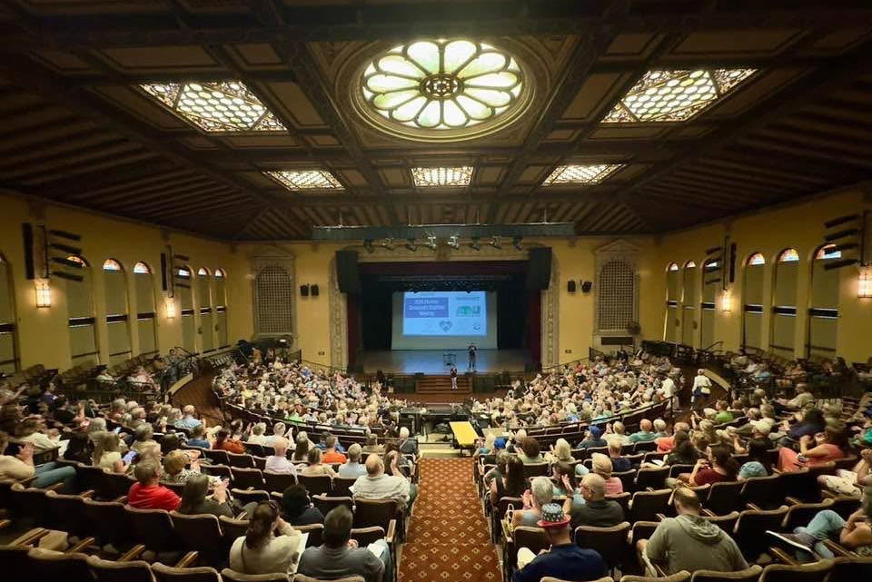 A picture taken from the back of the auditorium showing a packed house and speakers on the front stage
