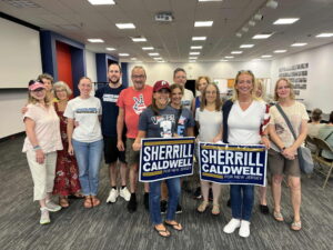 A group of people pose for a picture in a spacious office building. Some people are holding Sherril/Caldwell election signs and others wear shirts that say "Cooper River Indivisible"