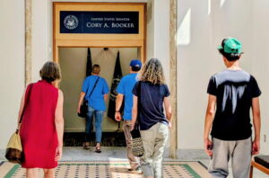 A group of people walk down a hall and into the offices of Cory Booker. We are viewing the people from behind and the sun is streaming in through a window on the right side of the picture