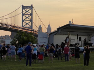 A picture of the Good Trouble Celebration in Camden. The sun is setting and the sky is orange. You can see the bridge and the city in the background as the people gather around a stage in the foreground