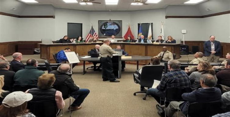 A townhall board room. The council people sit at a raised dias as the lesser functionaries sit at a table at ground level. A person speaks at a podium as the audience looks on