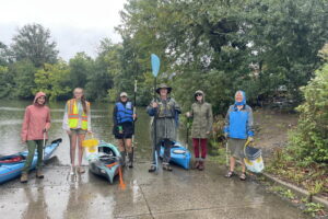 Six people pose for a picture at the boat ramp at Newton Lake in Oaklyn. Some hold kayak paddles and other grabbers for getting trash. All are dressed to deal with wet weather