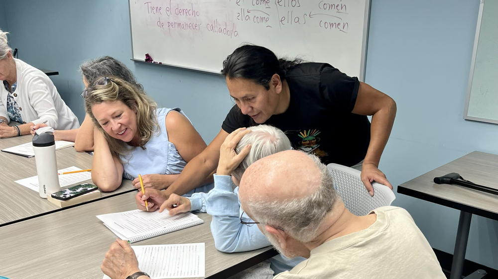 A group of people are sitting at a table hard at work on the Spanish Dialogs. The teacher leans over the table to help them with their work