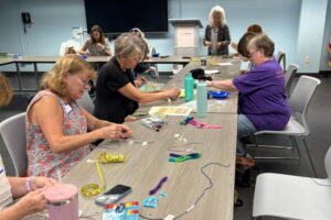 A group of people sit around a table in the Voorhees library making bracelets. On the table in front of them sit all the supplies that are needed.