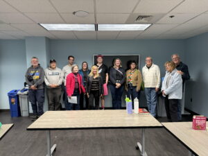 I group of people stand in front of a flat-screen tv on the wall of the Voorhees Library. There are 11 members of the book club and then the author of our most recent book. In front of the group are folding tables.