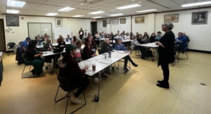 A crowded room with people sitting at tables listing to a presentation from the leader