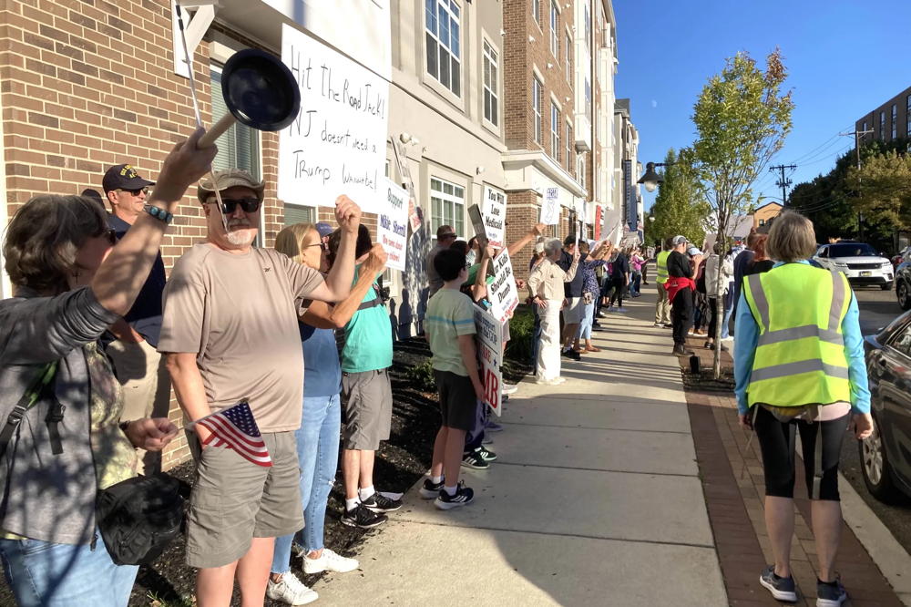 A line of protesters stand along the sidewalk on Haddon Ave holding signs. On the right side of the picture are safety marshals making sure that everyone is able to exercise their 1st amendment rights without fear