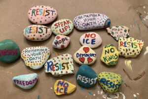 About a dozen painted rocks sit on a table. The rocks are colorful and have positive messages and protest messages on them. One says "No ICE" and one says "Hope"