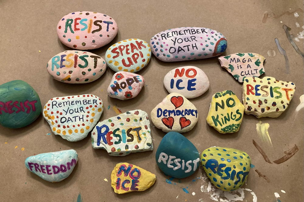 About a dozen painted rocks sit on a table. The rocks are colorful and have positive messages and protest messages on them. One says "No ICE" and one says "Hope"
