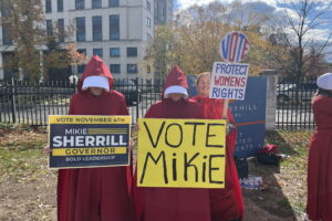 Three people wearing handmaids outfits stand in a line holding signs for Mikie Sherrill