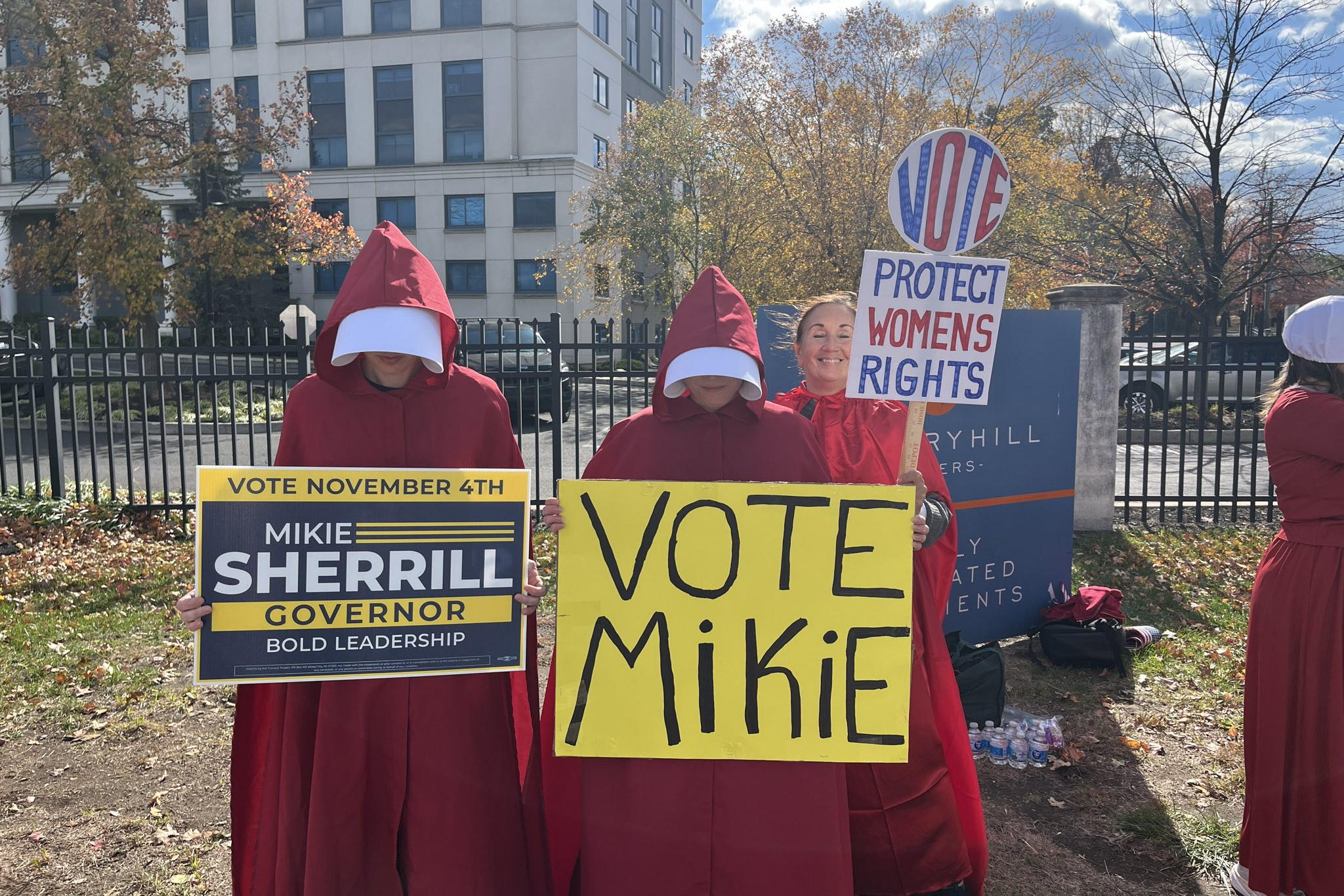 Three people wearing handmaids outfits stand in a line holding signs for Mikie Sherrill
