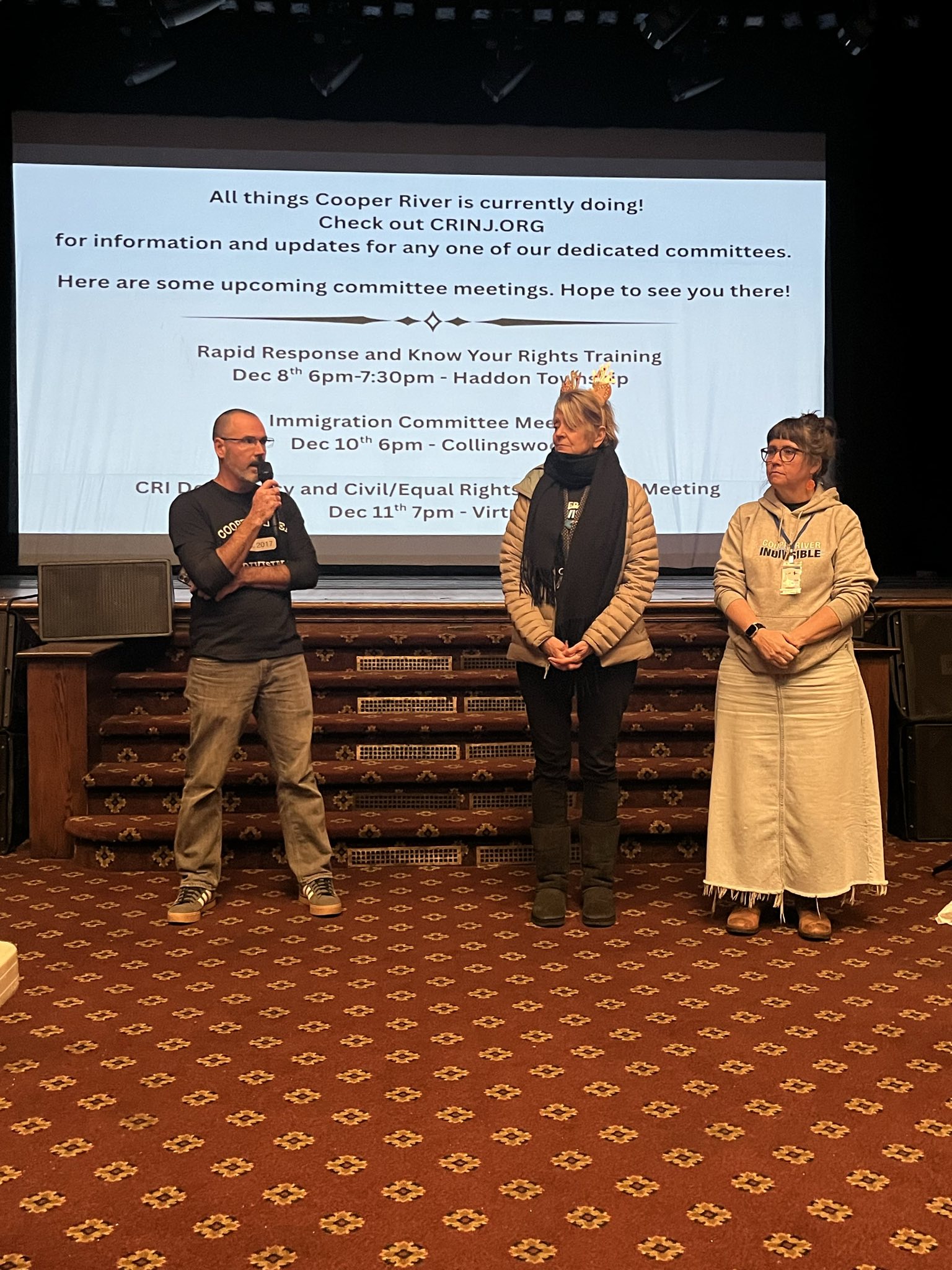 Three people stand in front of a stage. The one on the left is holding a microphone and speaking to the crowd. The two to the left are listening and waiting their turns. A list of events in behind them