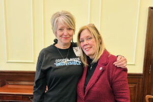 Two women stand in front of a light yellow wall. The one on the left is wearing a Cooper River Indivisible Shirt and the one on the right has a red blazer on