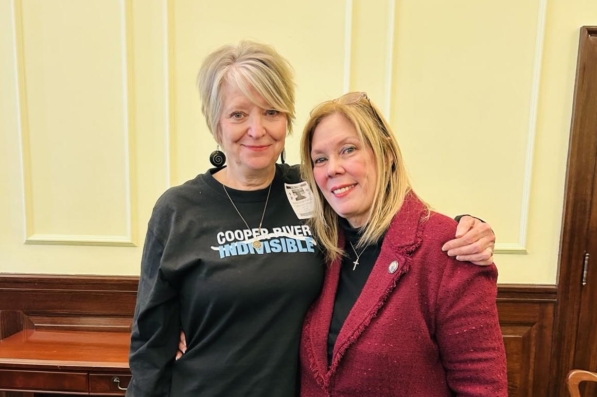 Two women stand in front of a light yellow wall. The one on the left is wearing a Cooper River Indivisible Shirt and the one on the right has a red blazer on