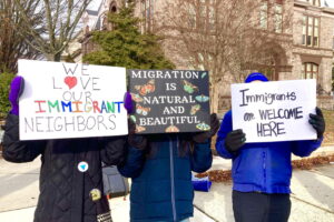 Three of 50 people from the protest along route 45 in Woodbury. The people stand on the sidewalk with an old stone building in the background. They hold signs in front of their faces to show support for immigrant rights.
