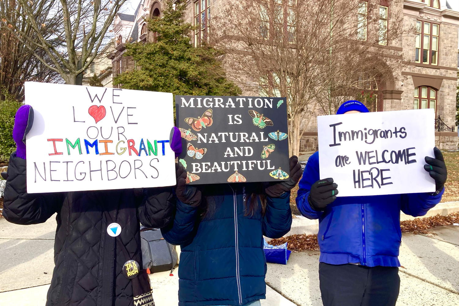 Three of 50 people from the protest along route 45 in Woodbury. The people stand on the sidewalk with an old stone building in the background. They hold signs in front of their faces to show support for immigrant rights.
