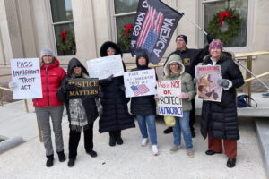 A group of cold people stand out front of the State House in Trenton holding signs and flags