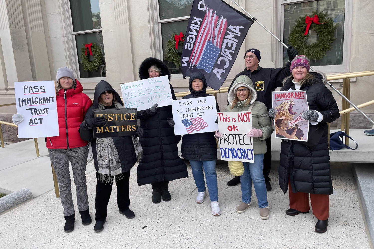 A group of cold people stand out front of the State House in Trenton holding signs and flags
