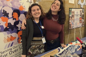 Two people stand behind tables at the SnowFlake shop . There are posters on the walls and goods on the tables