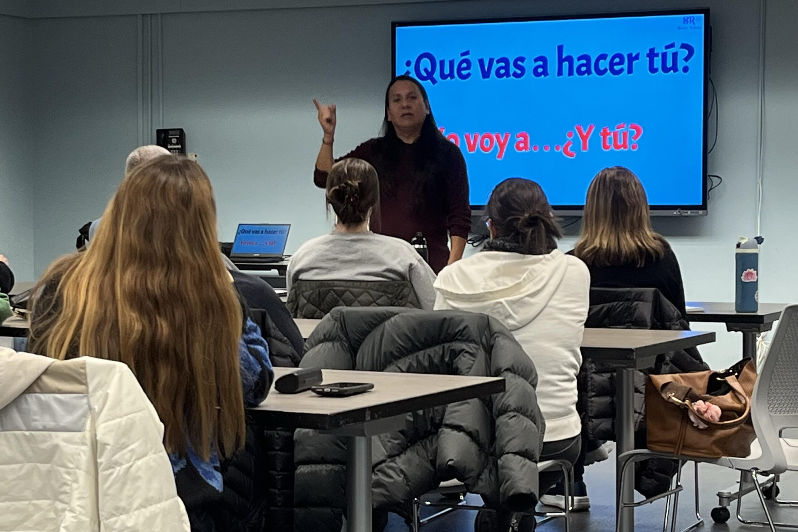 We are in the Voorhees library. A tv is in the background with a teacher standing in front of it. Students sit at tables in front of the teacher