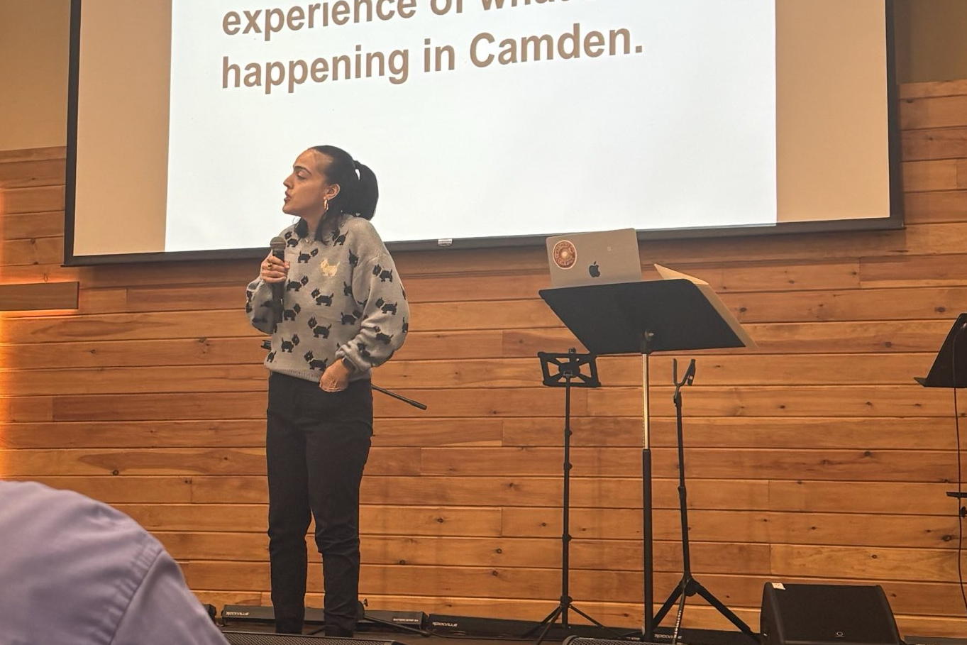 A teacher from Camden stands on stage with the microphone in hand. To the right is a music stand and in the background a description of the the talk