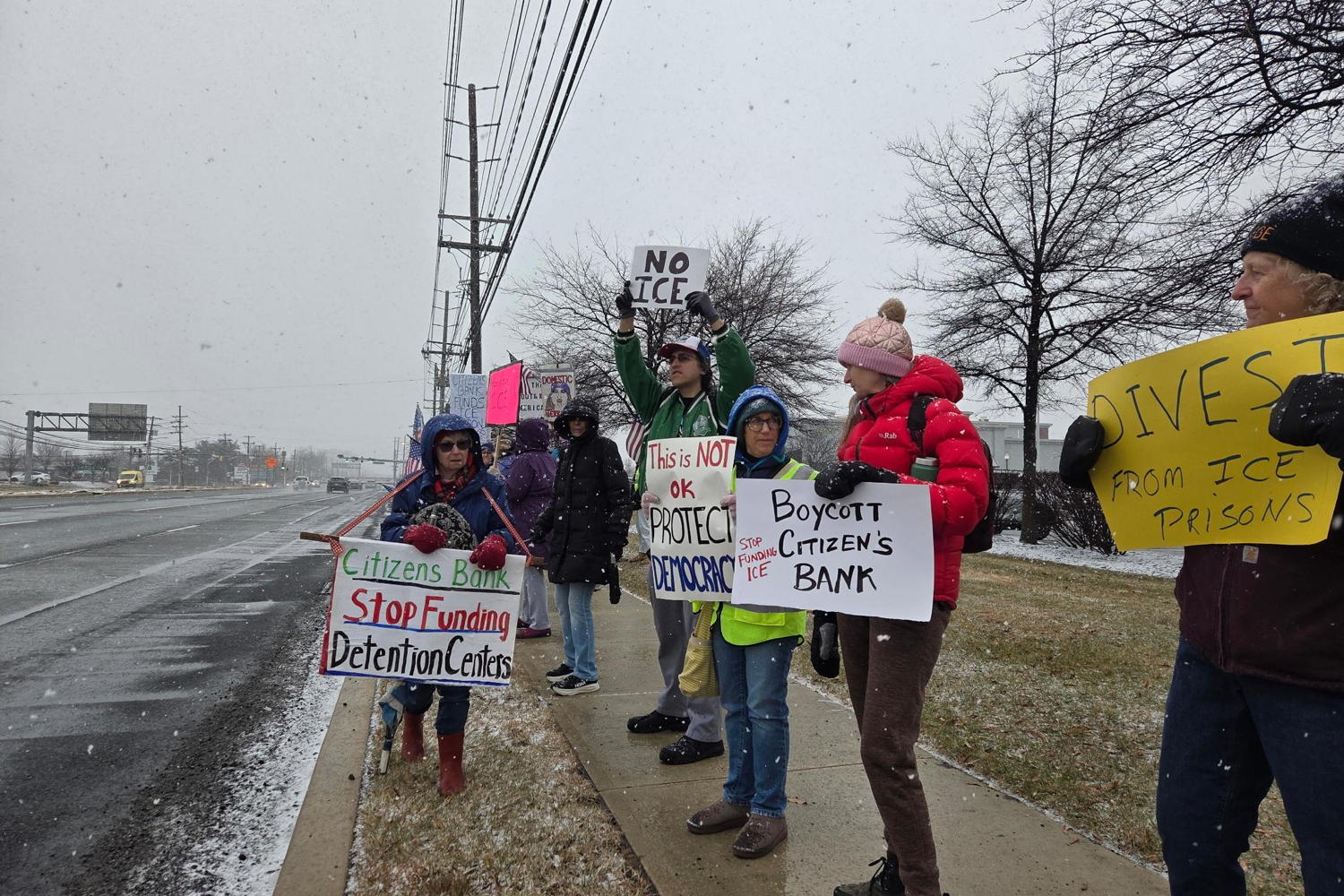 A group of people are protesting along the side of a very busy highway. Snow is falling around them and they are bundled up against the cold