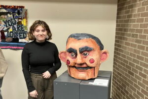 We are in an art show. A brick wall to the right and a display wall as the background. An artist stands next to their creation which is a head of Jack C made of paper mache