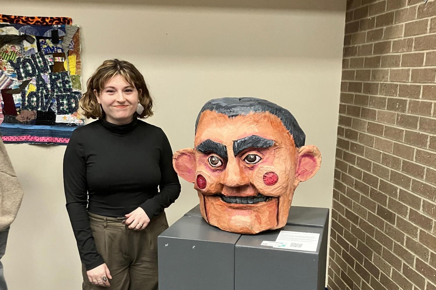 We are in an art show. A brick wall to the right and a display wall as the background. An artist stands next to their creation which is a head of Jack C made of paper mache