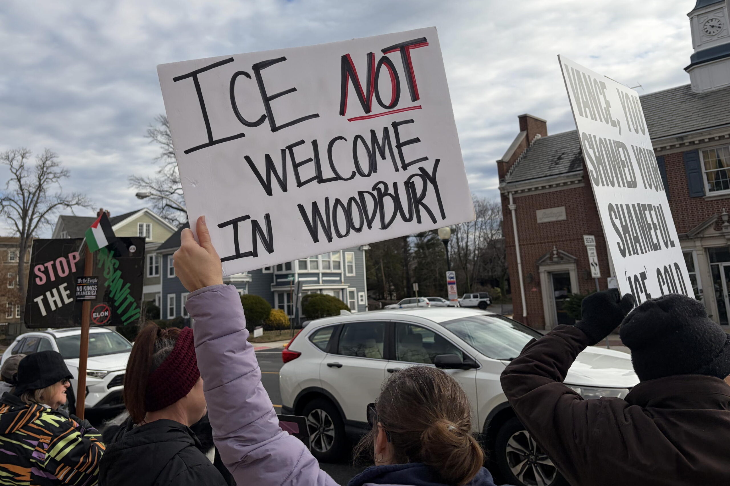 The sky above is mostly cloudy but with some hints of sunshine. The picture shows cars going by and people in the foreground holding signs. The most prominent sign reads "ICE NOT welcome in Woodbury"