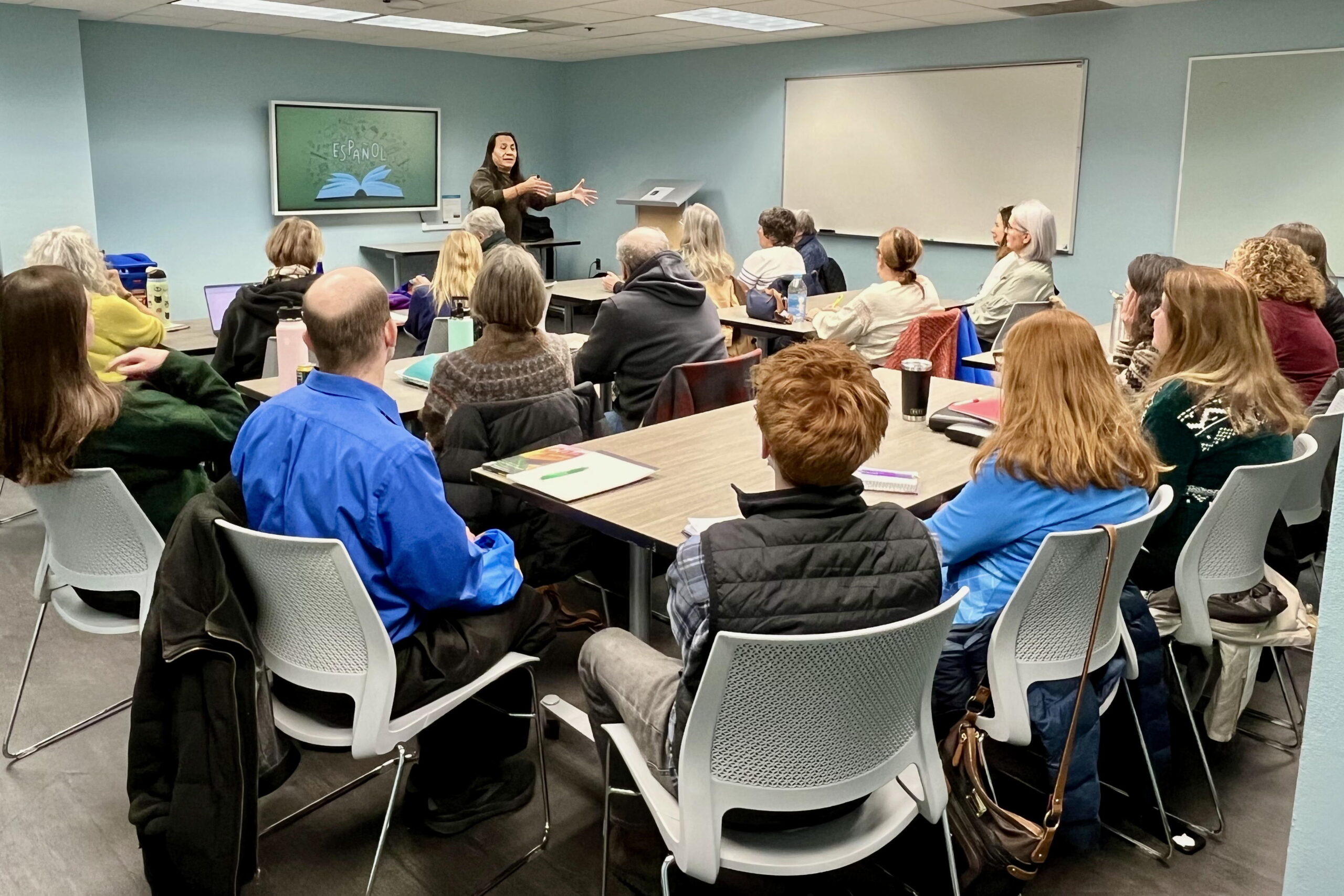 People sit around tables in a room in a library. The teacher is in the front of the room during the introductions