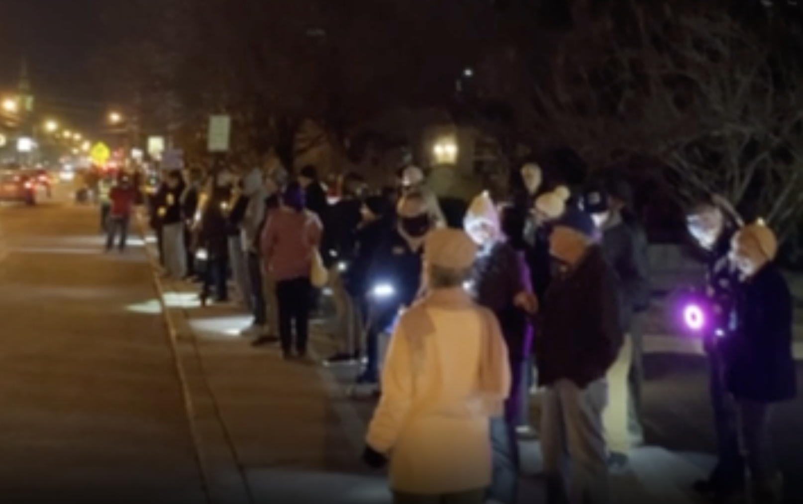 A shot looking east on Haddon Ave on a dark winter night. People line the south side of the road with candles and flashlights.