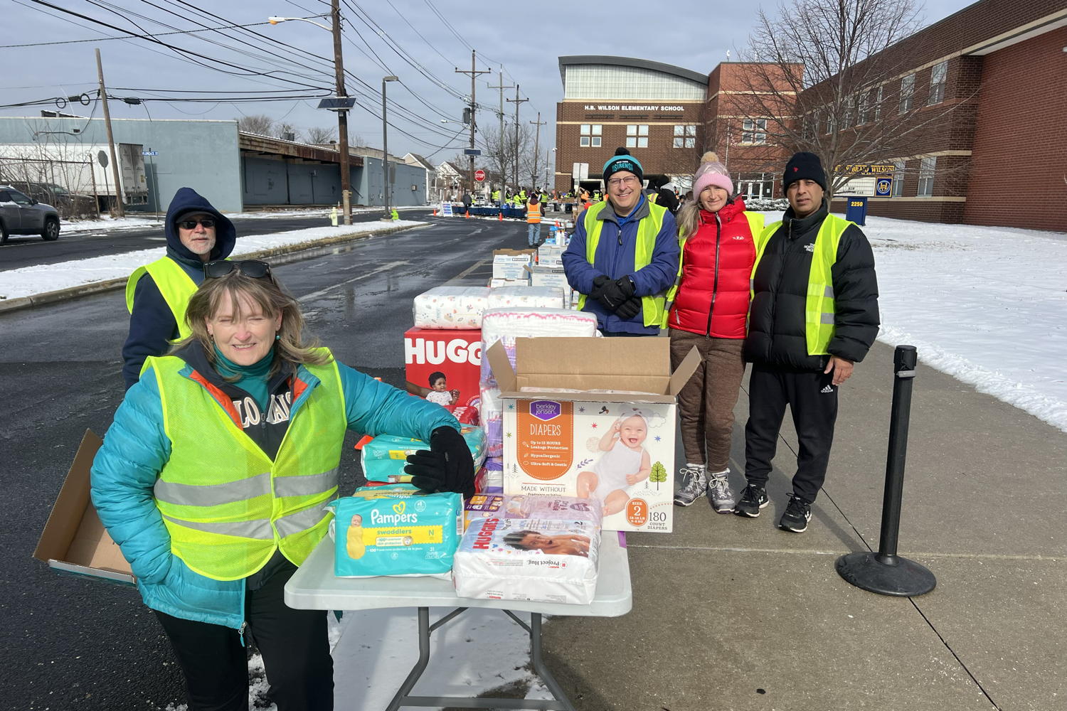 We are in front of H.B. Wilson Elementary School. Snow covers the grass and all roadways are wet. Members of CRI stand facing the camera with a table full of goods in front of us. Most of the items visible are diapers.