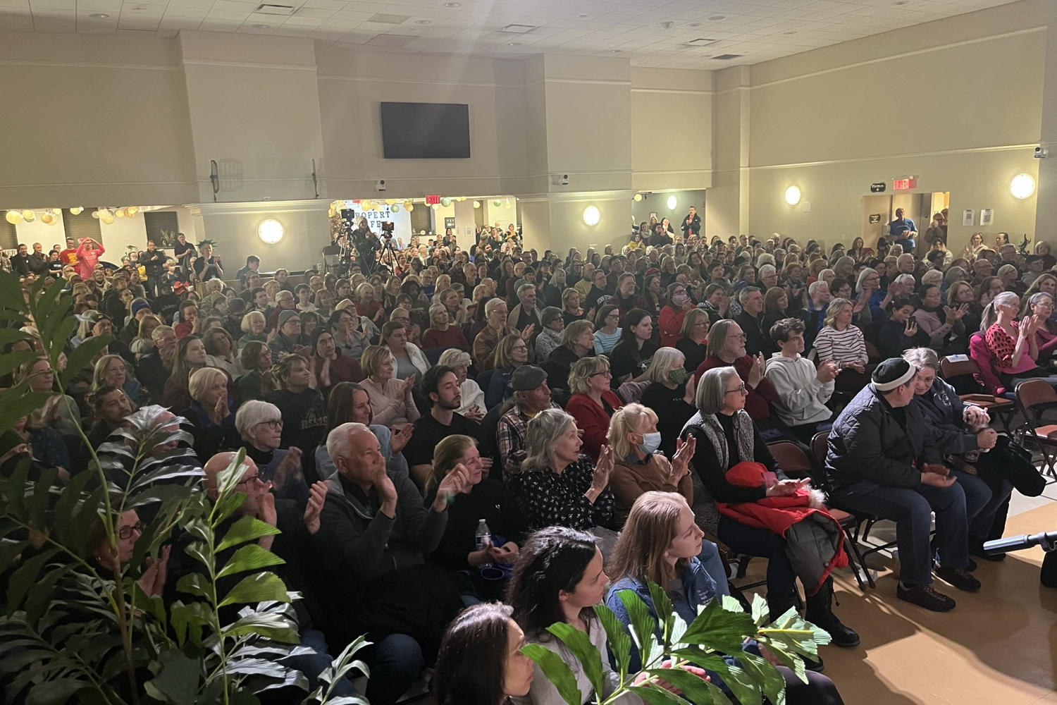 A view of the audience at our february general meeting taken from the stage. Everyone is clapping or listening intently. But one person, way in the back, in a pink shirt and pink hat is holding up their hands and making a heart symbol