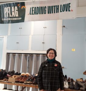 A picture of the clothing swap for PFlag. Above we see a banner that reads "Leading with Love" and then at waist level is a table filled with shoes. A work stand proudly in the front, off to the right