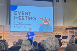A man in a blue shirt stand on stage speaking with a microphone. The slide welcoming people to the general meeting is in the background and the back of peoples' heads are in the foreground
