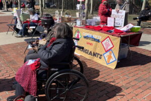 Members of Stitches for Solidarity sit and knit red hats. In the background is the table selling the hats