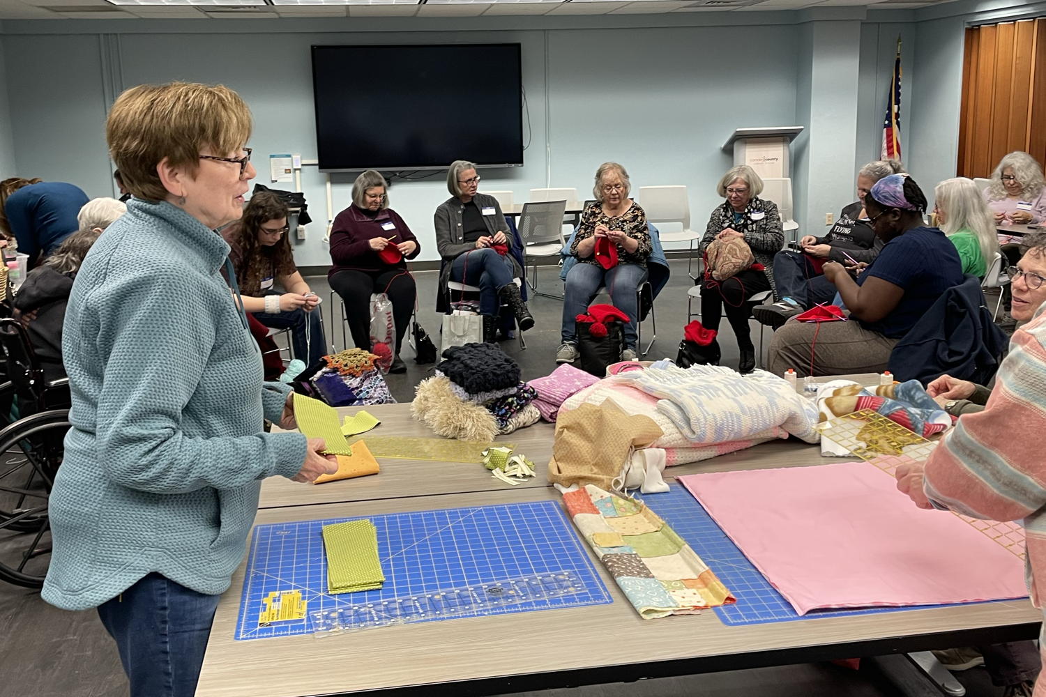 A room in the Voorhees library. In the foreground we have a table with a few women working on patterns. In the background we have sitting women working on making red hats