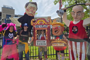 Our puppets pose for a picture. On the left we have a person in a furry suit. On the right is a person in a Jeff Bezo costume. In the middle is a ticket booth that says Pay to Play $1 Billion.