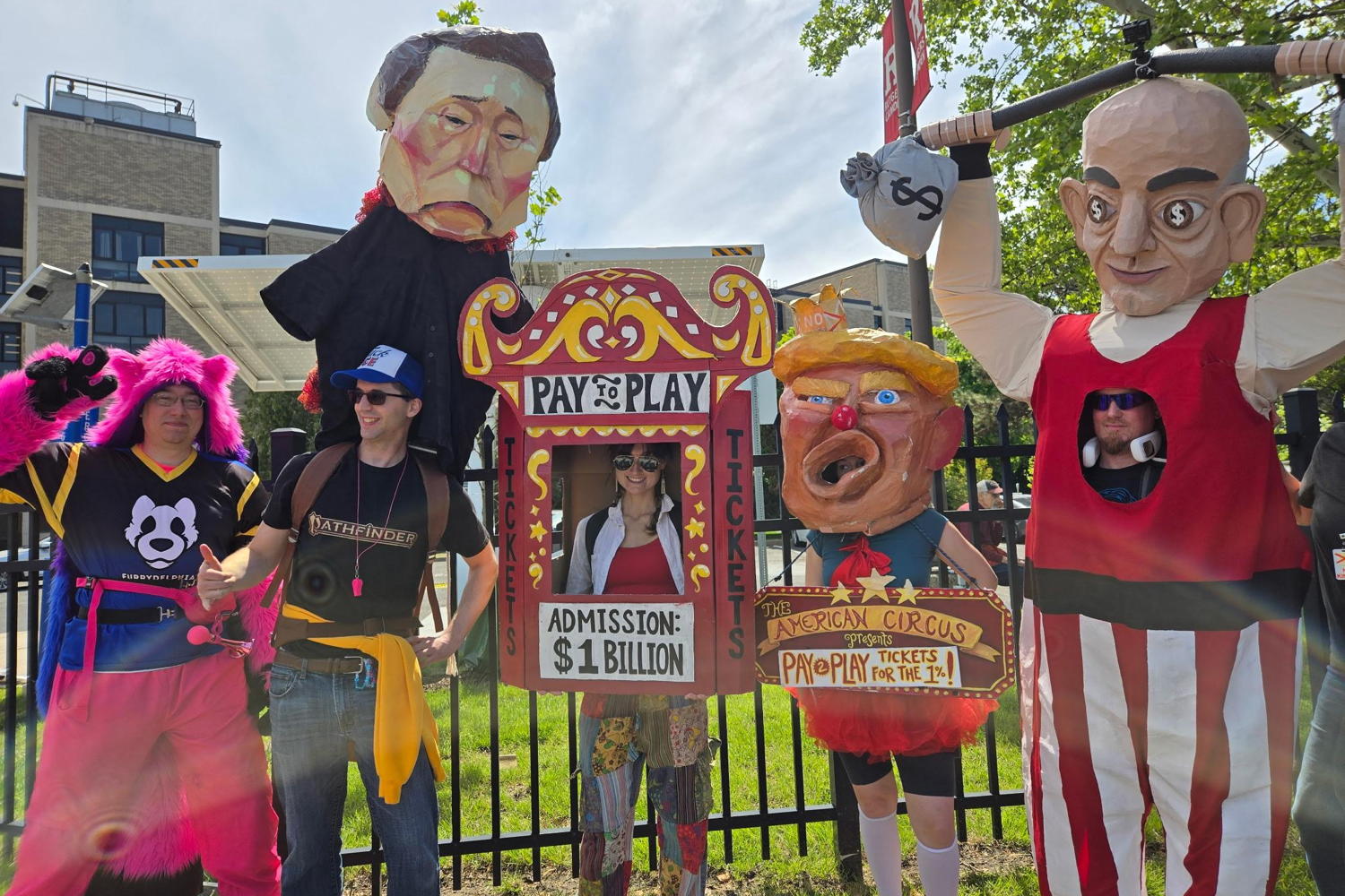 Our puppets pose for a picture. On the left we have a person in a furry suit. On the right is a person in a Jeff Bezo costume. In the middle is a ticket booth that says Pay to Play $1 Billion.