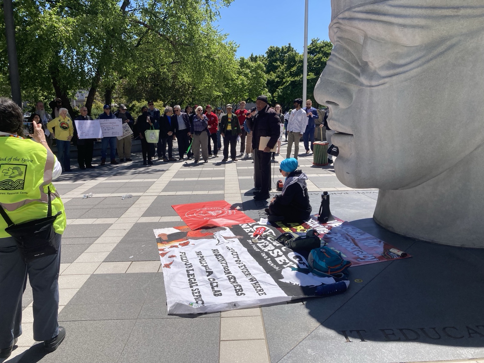 We are standing in the plaza in Newark. A giant concrete head is to the right. A person is giving a speech and people form a semi-circle around to listen. One person is sitting in the shade of the giant head.